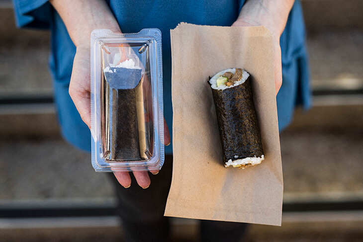 plastic packaged sushi roll next to sushi roll on paper bag